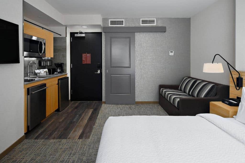 loft do TownePlace Suites by Marriott New York Manhattan/Times Square com uma cama de casal no canto inferior direito, ao lado de um sofá cama listrado que está de frente para uma cozinha compacta, equipada com microondas, máquina de lavar louças, frigobar, pia e cafeteira elétrica.