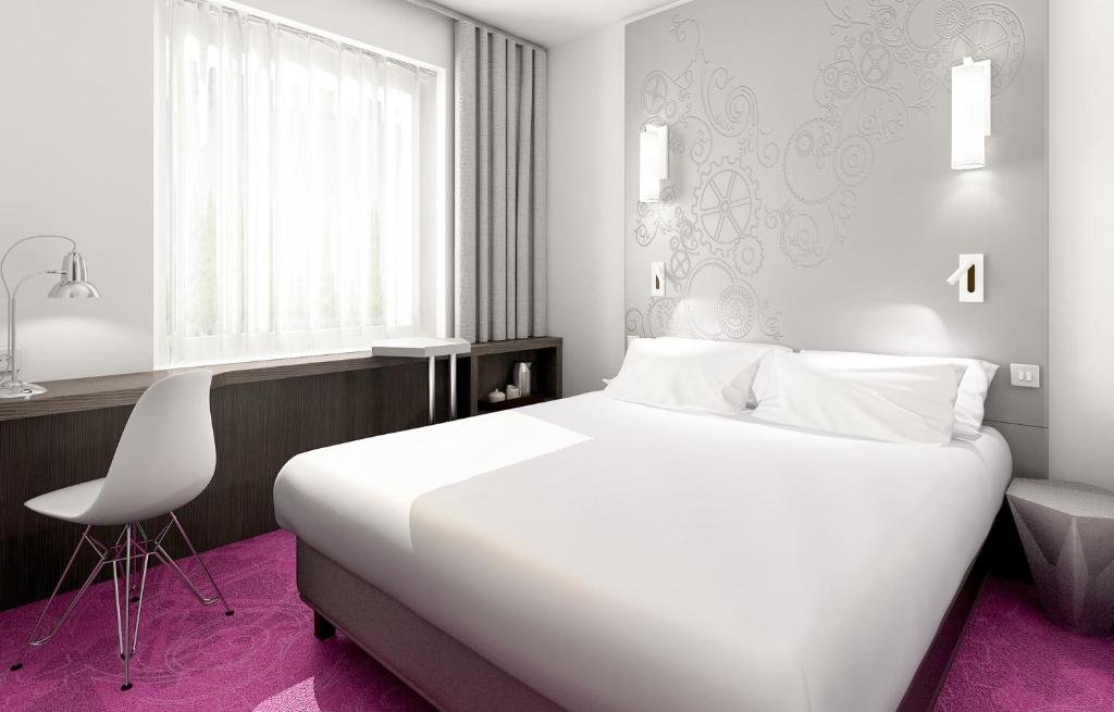 Foto de um VISIONAPARTMENTS Geneva Gare, para ilustrar o post de hotéis em Genebra. A cama king-size está no centro, encostada na parede à direita. As paredes são brancas e o carpete é roxo. Na esquerda, está a escrivaninha, que e estende pela parede inteira, e possui uma cadeira e abajur.