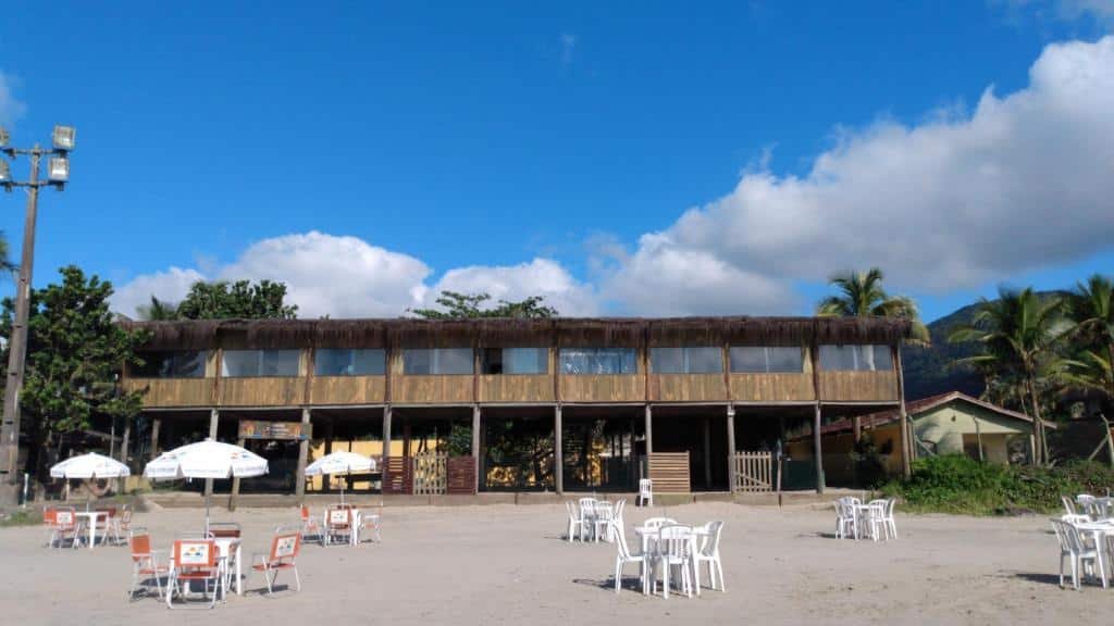 Frente do Suítes Pé na Areia. Mesas e cadeiras com guarda-sóis na areia da praia. Atrás a pousada.