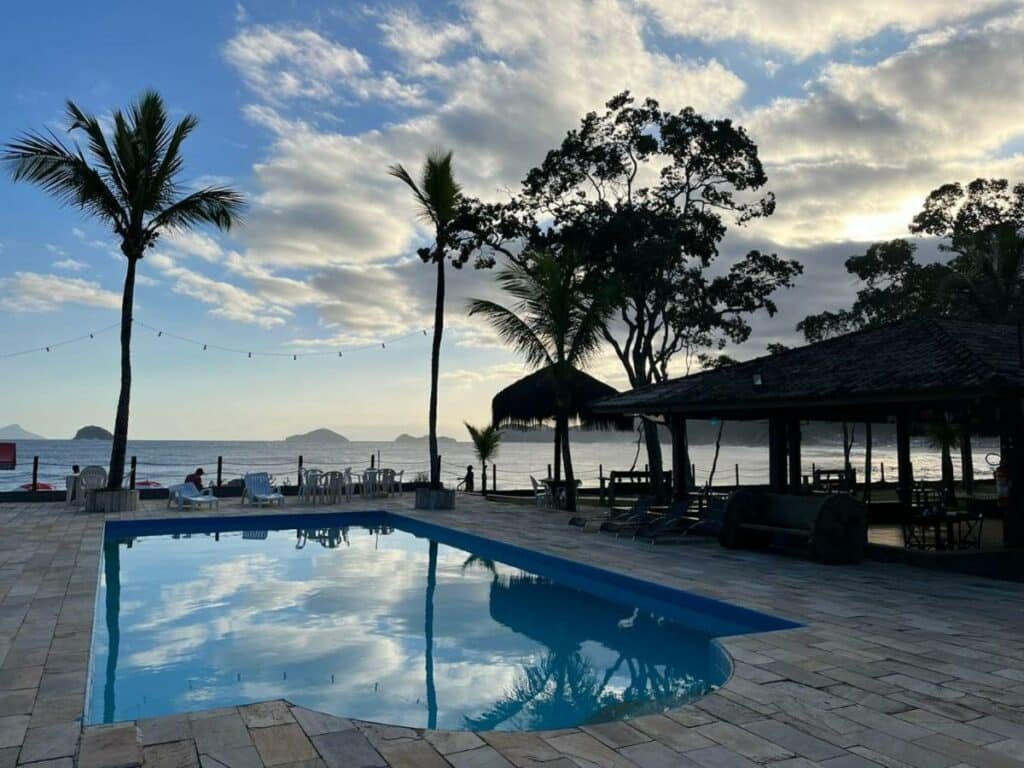 Piscina do Summit Beach Hotel Boiçucanga. Ela é grande, está perto de algumas cadeiras e de frente para a praia.