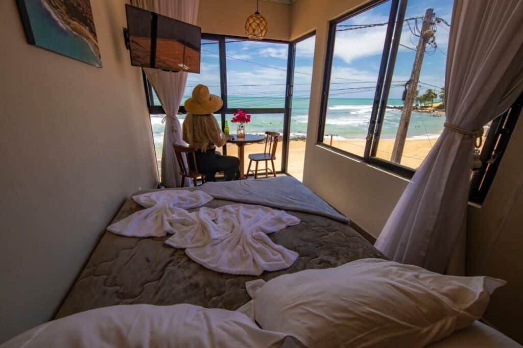 Quarto da pousada SuRFCoRe House. Uma cama ocupa todo o ambiente e está de frente para uma janela e uma porta com vista para a praia. Uma mesa está em frente a porta e uma tv está acima dela, de frente para a cama. Esta é mais uma foto para a lista de pousadas em Baía Formosa, RN.