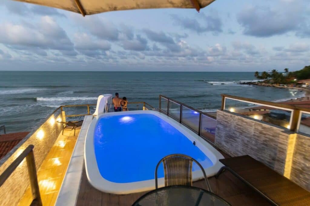 Piscina da pousada SuRFCoRe House. Ela possui formato retangular com as bordar arredondadas, uma pequena cascata de água e está de frente para o mar em uma área elevada e iluminada. Atrás dela é possíver ver também uma mesa com cadeira.