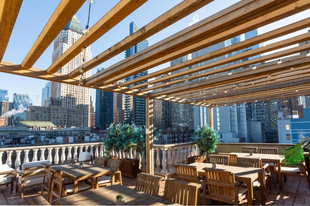 terraço do Hotel Scherman com várias mesas e cadeiras de madeira embaixo de várias vigas de madeira equipadas com luzinhas para decoração. Ao fundo é possível ver vários prédios de Manhattan, incluindo o famoso Empire State Building.