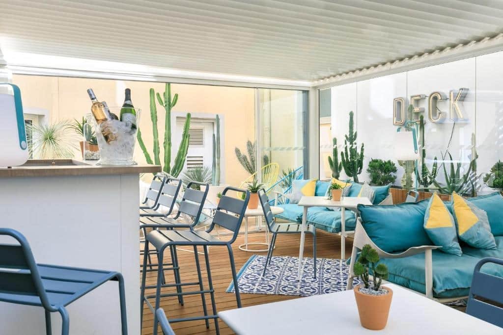 Bar do The Deck Hotel by Happyculture. No lado esquerdo está o balcão do bar com um balde de gelo com vinhos e banquetas na frente. No lado direito há alguns sofás, mesas, cadeiras e plantas decorativas.