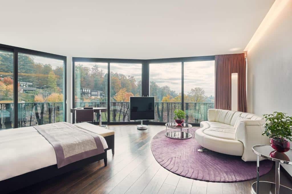Suíte Júnior deluxe do The Dolder Grand, um dos hotéis em Zurique, de 54 m², com janelas fora a fora de vidro mostrando de maneira panorâmica a natureza em volta, composta por árvores e folhas amarelas, sendo dentro do quarto uma cama de casal, uma TV quadrada, uma mesa redonda com uma cadeira e um sofá branco no formato oval