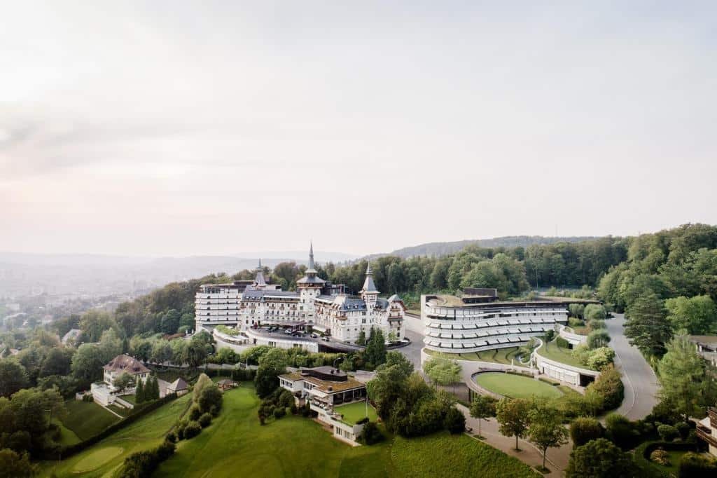 Vista ampla e panorâmica do The Dolder Grand, com uma arquitetura similar a um castelo, com algumas torres e vários edifícios brancos, além de bastante verde composto por uma natureza vasta nas redondezas