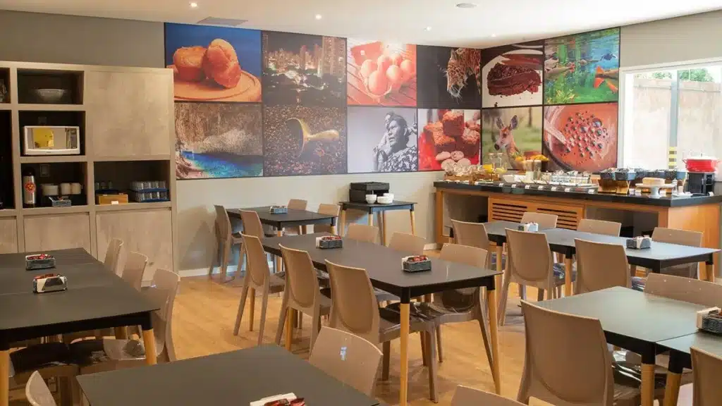 Área de refeições com várias mesas e cadeiras, parede decorada, armário com eletrodomésticos e coisas de cozinha. No outro canto há uma mesa com café da manhã.