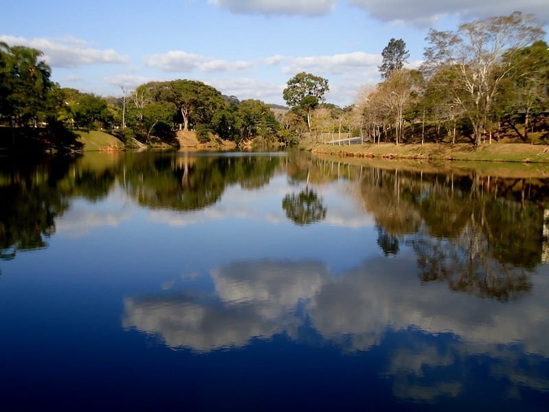 Imagem de um lago na Universidade Federal de Viçosa. A lago é grande e está refletindo as nuvens no céu. No fundo estão diversas árvores. Imagem para ilustrar o post hotéis em Viçosa.