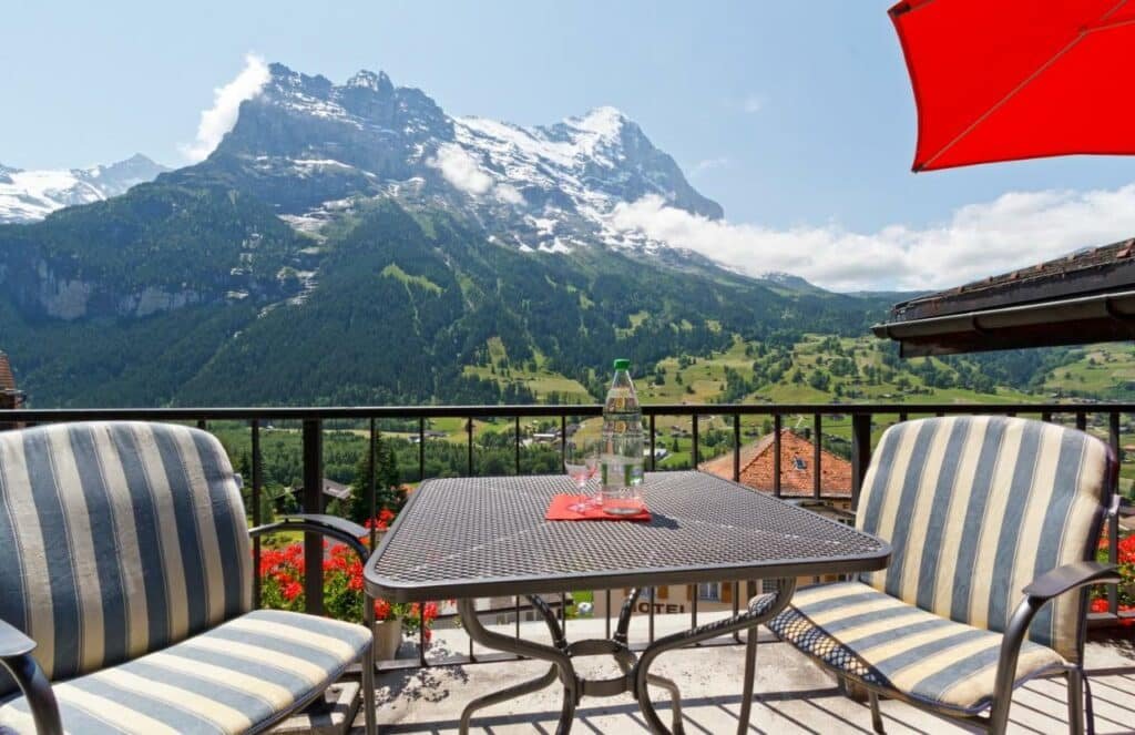 Imagem da varanda do Derby Swiss Quality Hotel com uma mesa quadra e duas cadeiras com vista para as montanhas.
