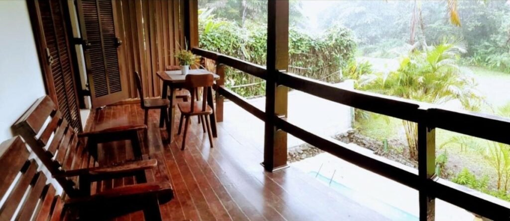 Varanda do Flat na Jureia – 30 MT da Praia durante o dia com cadeiras de madeira do lado esquerdo da imagem a frente e ao fundo uma mesa de madeira com três cadeiras.