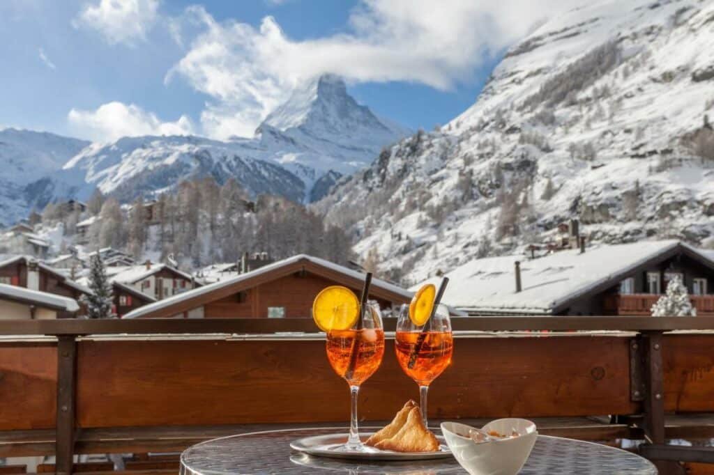 Varanda do Hotel Eden Wellness durante o dia com uma mesa redonda com duas taças com drinks, pães em cima com vista para os telhados e montanhas da cidade.