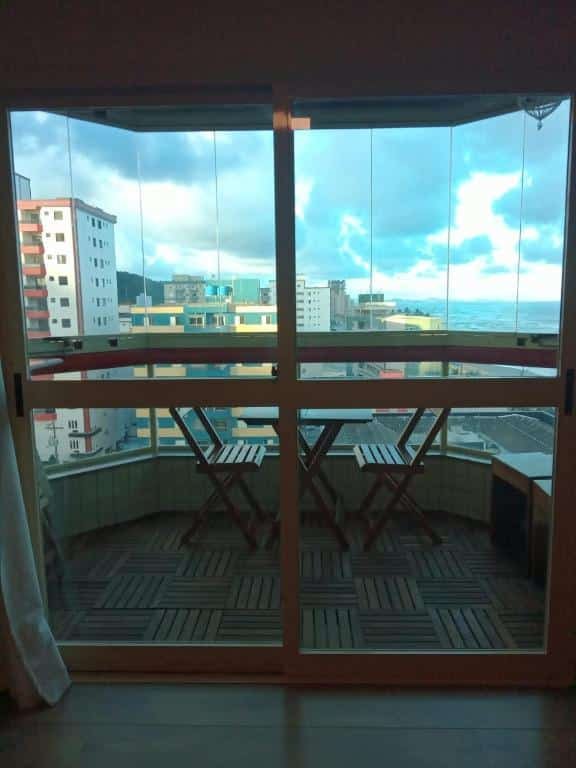 Porta de vidro que vai para varanda de apartamento com chão, mesa e cadeiras de madeira e vista para o mar e outros prédios.
