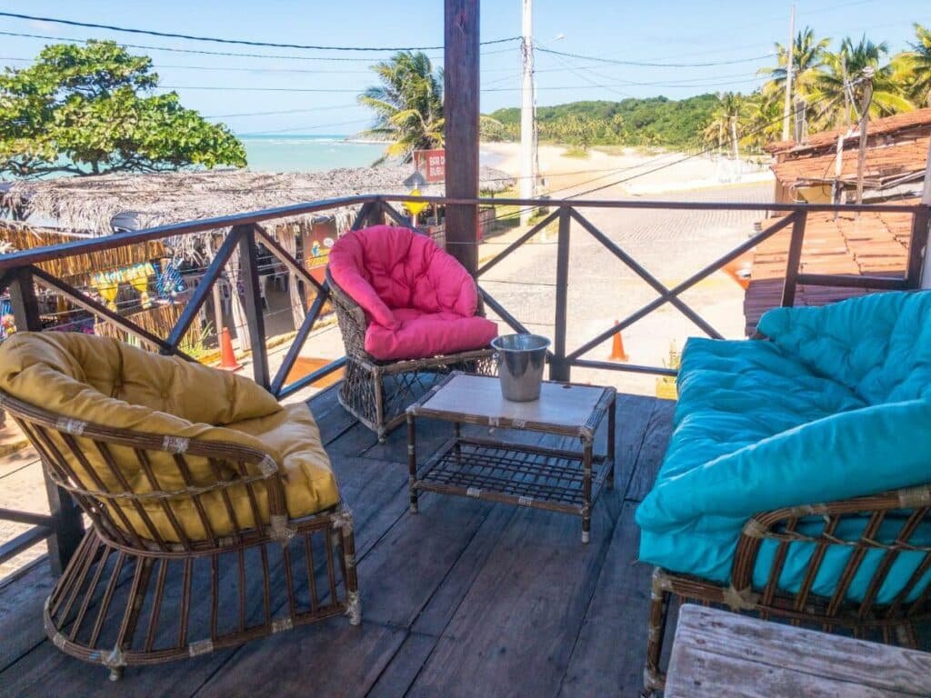 Varanda da casa Verano House BF com três cadeiras acolchoadas dispostas em formato circular e com uma mesinha no meio. Ao fundo é possível ver a rua, um quiosque e a praia. Esta é mais uma das opções na lista de pousadas em Baía Formosa.
