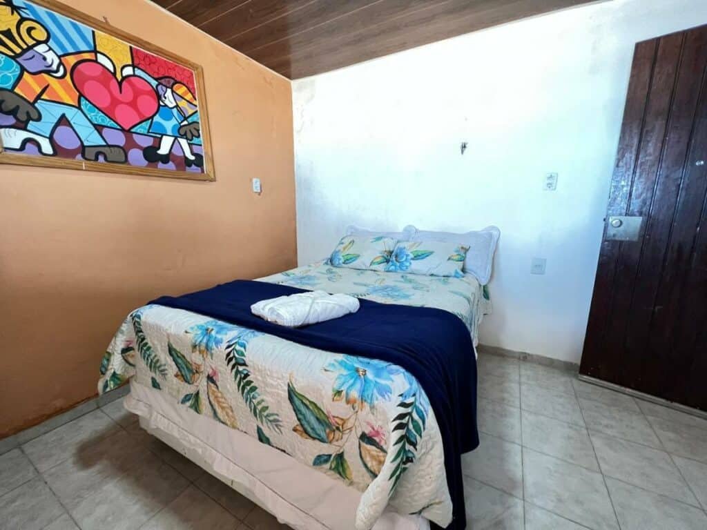 Quarto da Verano House BF. Uma cama de casal está quase encostada na parede do lado esquerdo. Ao lado direito há uma porta.