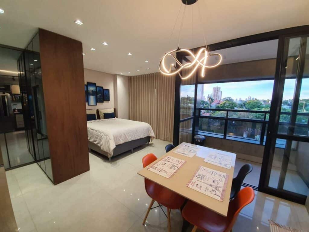Apartamento com uma cama de casal, armário ao lado e uma luminária iluminando uma mesa com cadeiras. Há também uma varanda com vista para a cidade.