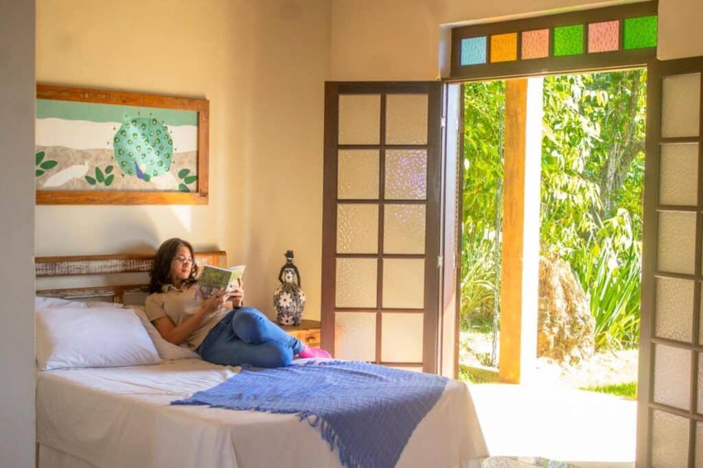 Uma pessoa lendo um livro em uma cama perto de portas francesas abertas, com uma cabeceira pintada de cores vivas e vista para a vegetação do lado de fora na Vila Chico.