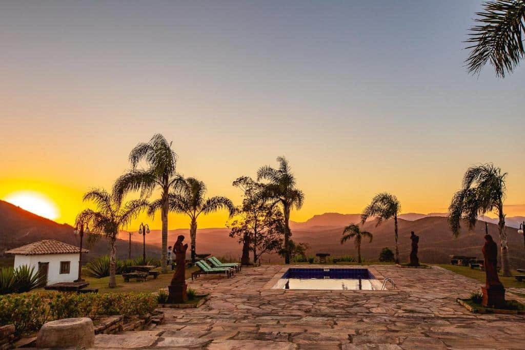 A imagem mostra uma área externa com uma piscina retangular, rodeada por espreguiçadeiras e palmeiras, durante o pôr do sol. O céu está tingido de tons alaranjados e amarelos, enquanto o sol se põe por trás das montanhas ao fundo. Estátuas decorativas estão dispostas ao redor da área de lazer, e o chão é pavimentado com pedras naturais. A paisagem é serena, com montanhas e vegetação ao longe, criando um cenário tranquilo e ideal para relaxar. Uma pequena casa branca com telhado de telhas vermelhas está visível à esquerda. Esse é o Hotel Vila Relicário, um dos hotéis-fazenda em Minas Gerais.