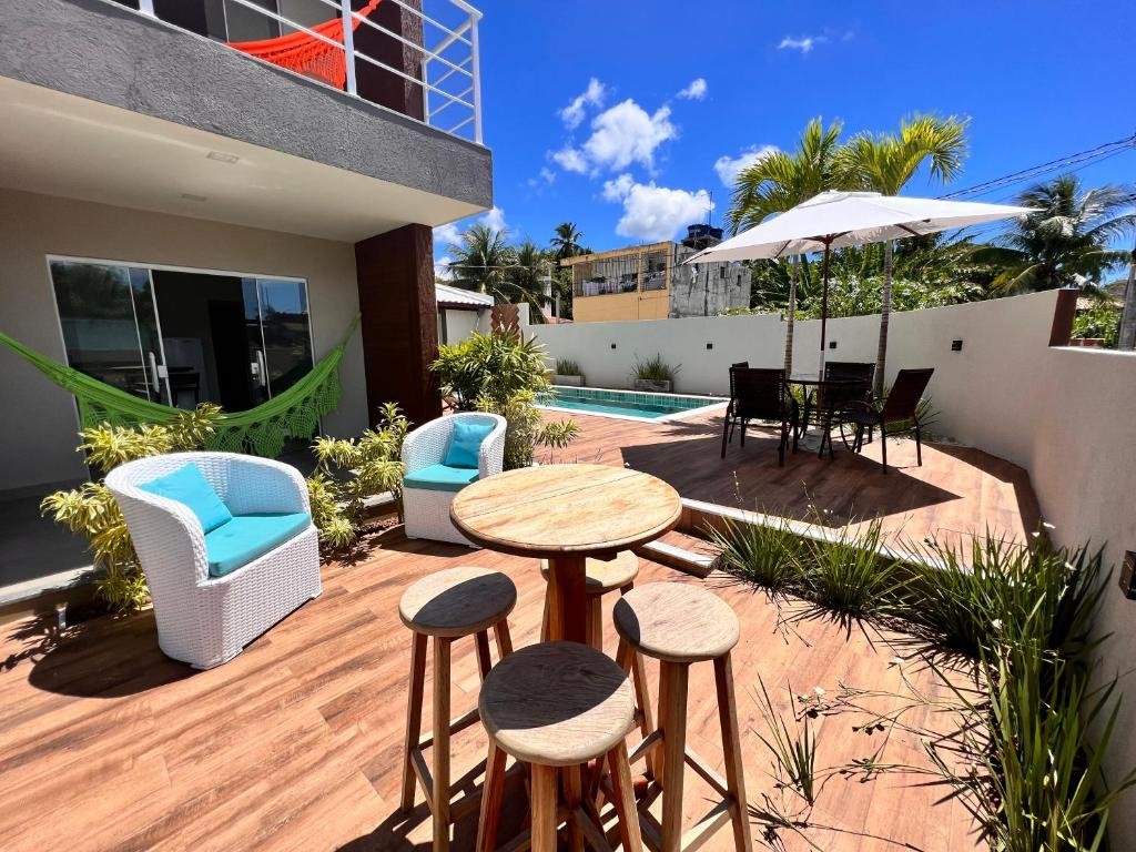 Área externa da VilaMar Jacuípe em Camaçari. Uma mesa redonda na frente com quatro bancos de madeira, do lado esquerdo os quartos, uma rede e duas poltronas com almofadas, no lado direito um guarda-sol com cadeiras, uma mesa e no funda a piscina.