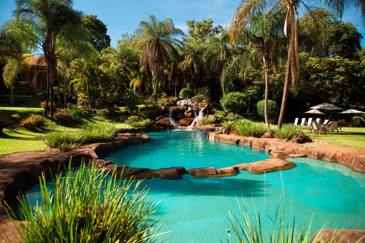 Piscina ao ar livre no Ville Real Hotel, um dos hotéis fazenda em Minas Gerais, cercada por vegetação tropical com espreguiçadeiras e guarda-sóis ao fundo, situada em um jardim exuberante.