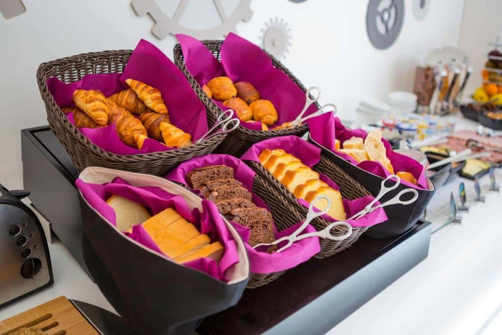 Buffet do VISIONAPARTMENTS Geneva Mont-Blanc. Vemos uma bancada branca com muitas opções de comida. Em foco, vemos várias cestas com guardanapos roxos com pães e massas dentro.