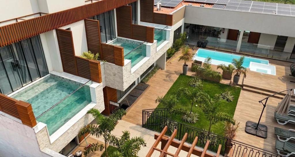 A imagem mostra uma vista aérea da Pousada Villas em Bonito. O complexo apresenta uma arquitetura moderna com linhas retas e acabamentos em madeira e pedra. Há pequenas piscinas privativas em frente a cada suíte, protegidas por divisórias de madeira e vidro. No centro do pátio, há uma área verde com plantas e palmeiras, criando um ambiente tropical e relaxante. Ao fundo, é possível ver uma piscina maior compartilhada, acompanhada de uma banheira de hidro. O espaço externo é bem cuidado, com um deck de madeira e espreguiçadeiras para os hóspedes relaxarem.