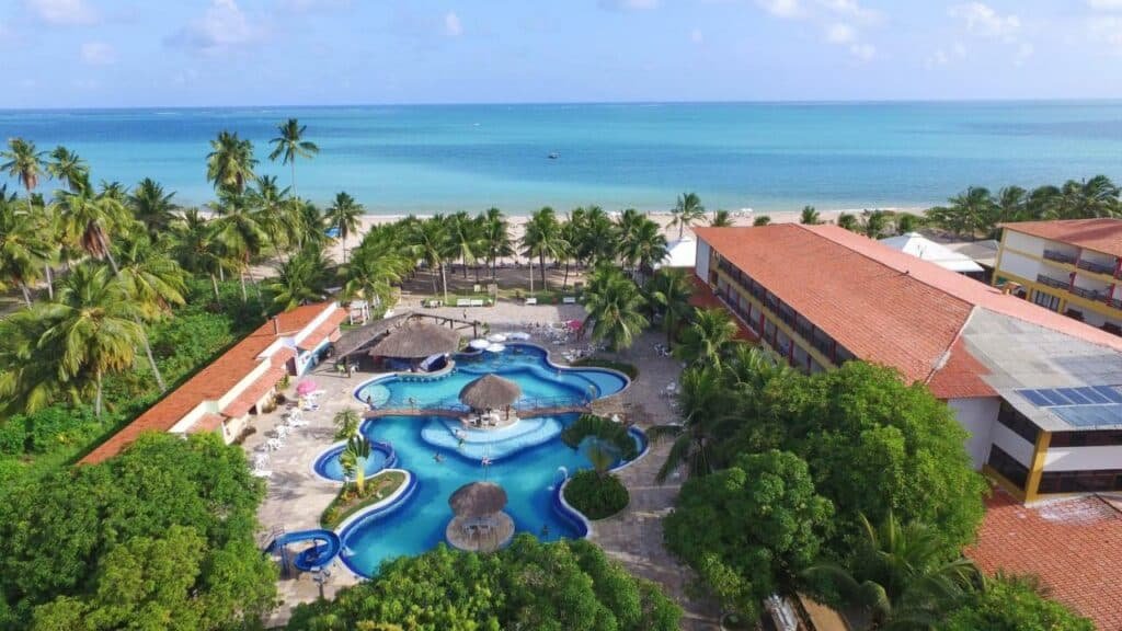 A imagem mostra uma vista aérea do Hotel Praia Dourada, um resort em Maragogi à beira-mar. O resort é cercado por abundantes palmeiras e está situado diretamente em frente a uma praia de areia com o oceano de águas claras ao fundo. No centro da imagem, há uma grande piscina em forma de lagoa com várias ilhas e uma área com toboáguas. As piscinas são rodeadas por espreguiçadeiras brancas dispostas sobre o pavimento. À esquerda, há edifícios de um andar com telhados vermelhos que parecem ser parte do complexo do resort. O ambiente é tropical e convida ao lazer, oferecendo uma mistura de vegetação densa, estruturas de resort confortáveis e a vasta extensão azul do oceano ao fundo.
