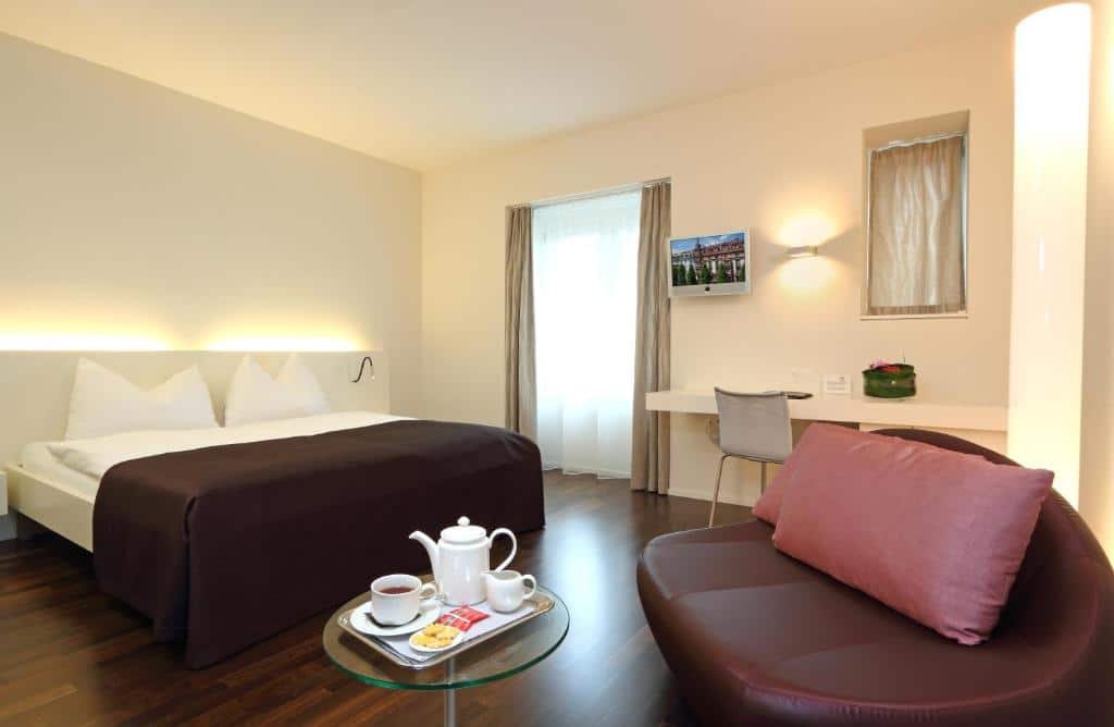 Foto do Waldstätterhof Swiss Quality Hotel, para representar o post de hotéis em Lucerna. A cama box de casal está na esquerda. Na direita, vemos uma poltrona grande de couro com travesseiros e uma pequena mesa à sua frente, essa que possui um bule de chá e xícaras . Atrás da poltrona, encostada na parede do fundo do quarto, está uma escrivaninha com cadeira e TV. Do seu lado esquerdo há uma janela.