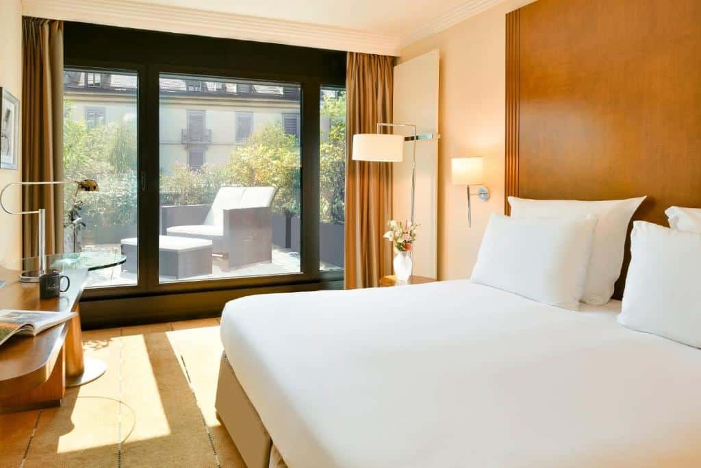Foto de um quarto do Warwick Geneva, para ilustrar o post de hotéis em Genebra. A cama king-size está no canto direito da imagem. Do outro lado, na parede do fundo, está a janela com vista para a varanda. Na frente da cama, há uma mesa e escrivaninha com revista caneca e abajur.