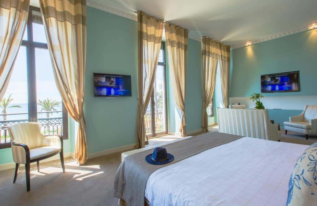 Quarto do Westminster Hotel & Spa Nice. A cama está centralizada no quarto e em frente há duas janelas enormes, uma porta no centro que dá acesso a varanda, uma TV e uma cadeira. No lado direito do quarto há uma pequena sala de estar com um sofá, duas poltronas, mesa de centro e outra TV. Imagem para ilustrar o post hotéis em Nice.