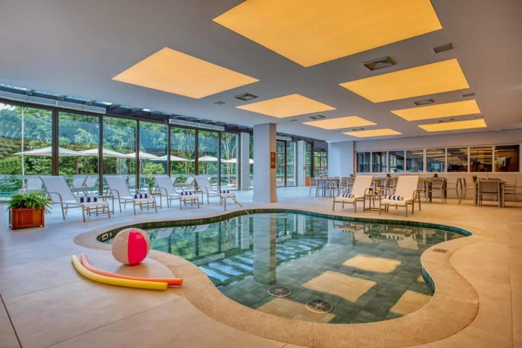 Piscina coberta e cercada por espreguiçadeiras e portas de vidro com direção para as árvores no Wyndham Gramado Termas Resort & Spa para representar airbnb em Gramado