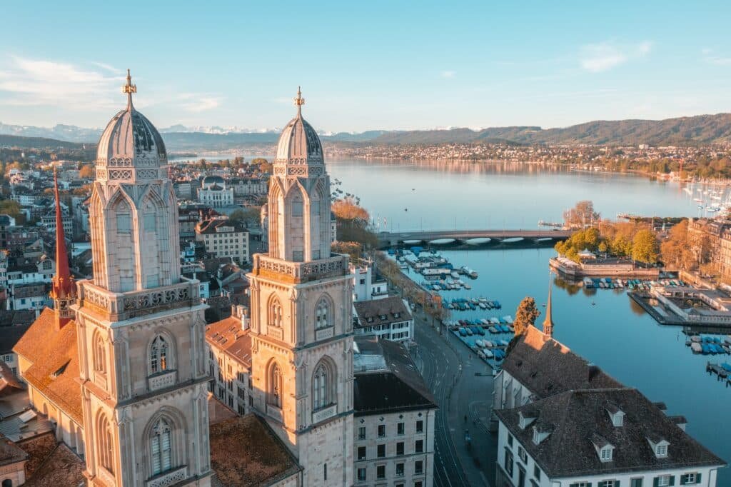 Vista das duas torres da Igreja de Grossmünster, com rio cristalino em volta e paisagem com montanhas no horizonte e casas